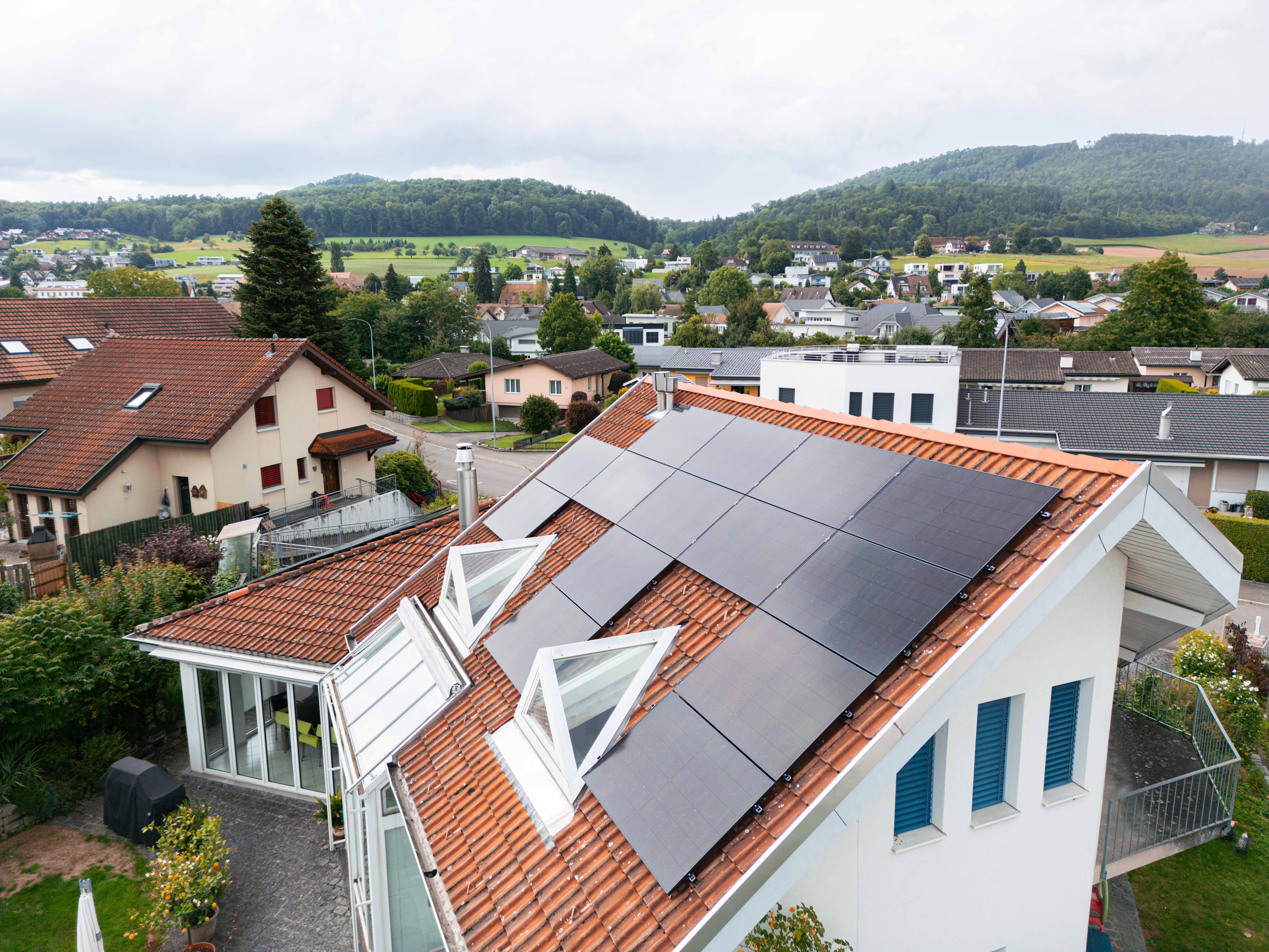 Solarmodule auf einem Ziegeldach neben zwei geöffneten Dachfenstern, dahinter das Dorf